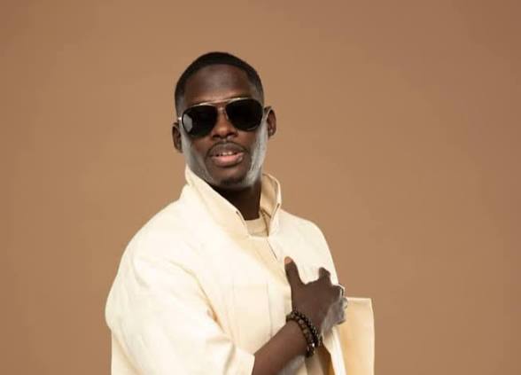 Momo Dieng pose en veste ivoire et lunettes de soleil devant un fond beige.