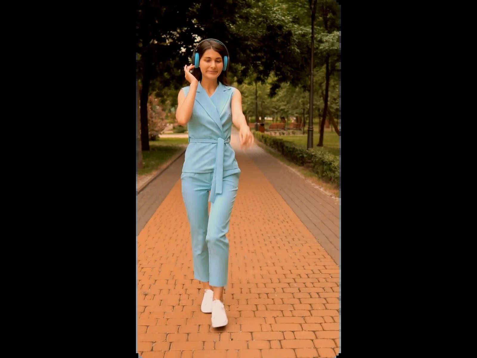 Femme au casque audio bleu marchant en tenue bleu clair sur une allée de briques dans un parc verdoyant